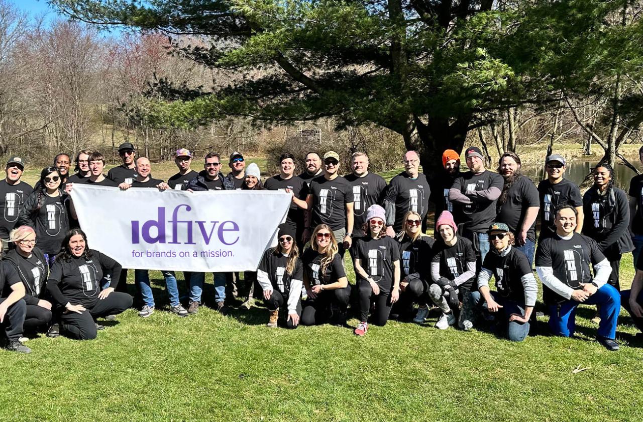A large group of idfive employees pose outdoors looking at the camera, holding up an idfive banner.